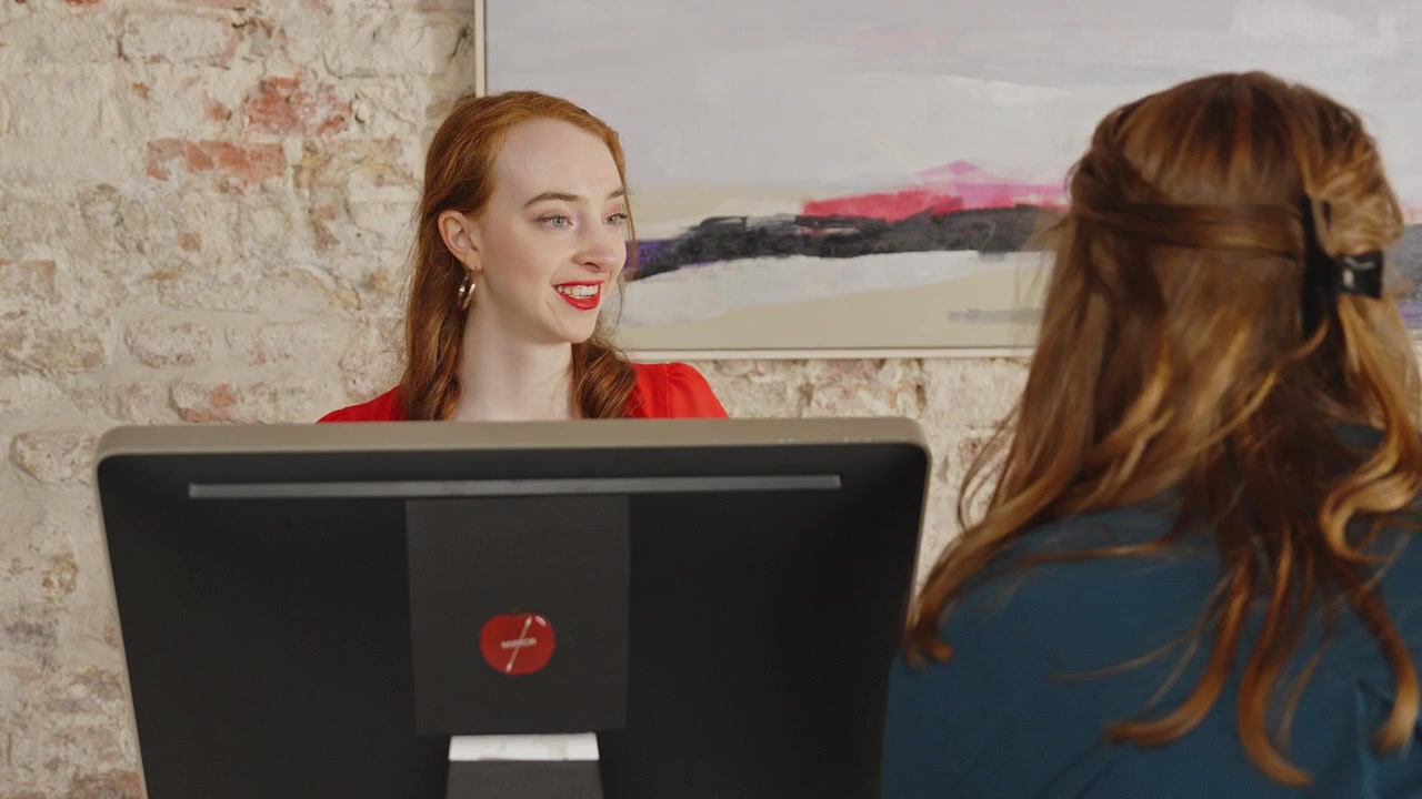 Una giovane receptionist reagisce scioccata quando un visitatore cerca di intimidirla. Lei cerca di disinnescare la situazione e sorride.