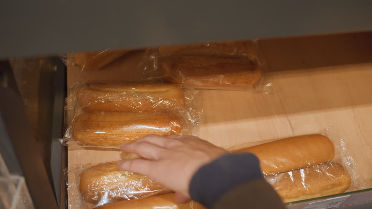 packaged bread loaves on shelf, clerk arranging wrapped buns in wooden drawer, plastic wrapping reflecting warm store lights, hands placing packages into neat rows, morning restock sequence