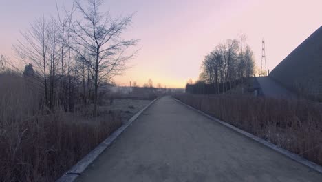 Die-Drohne-Folgt-Dem-Fußweg-Durch-Den-Park-Und-Fliegt-Nach-Oben,-Enthüllt-Einen-Teich-In-Der-Nähe-Und-Einen-Zug,-Der-Bei-Sonnenaufgang-Vorbeifährt