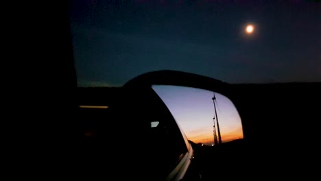 Long-row-of-wind-turbines-seen-through-rear-view-mirror-with-hazy-moon-in-the-background-during-dusk-while-driving