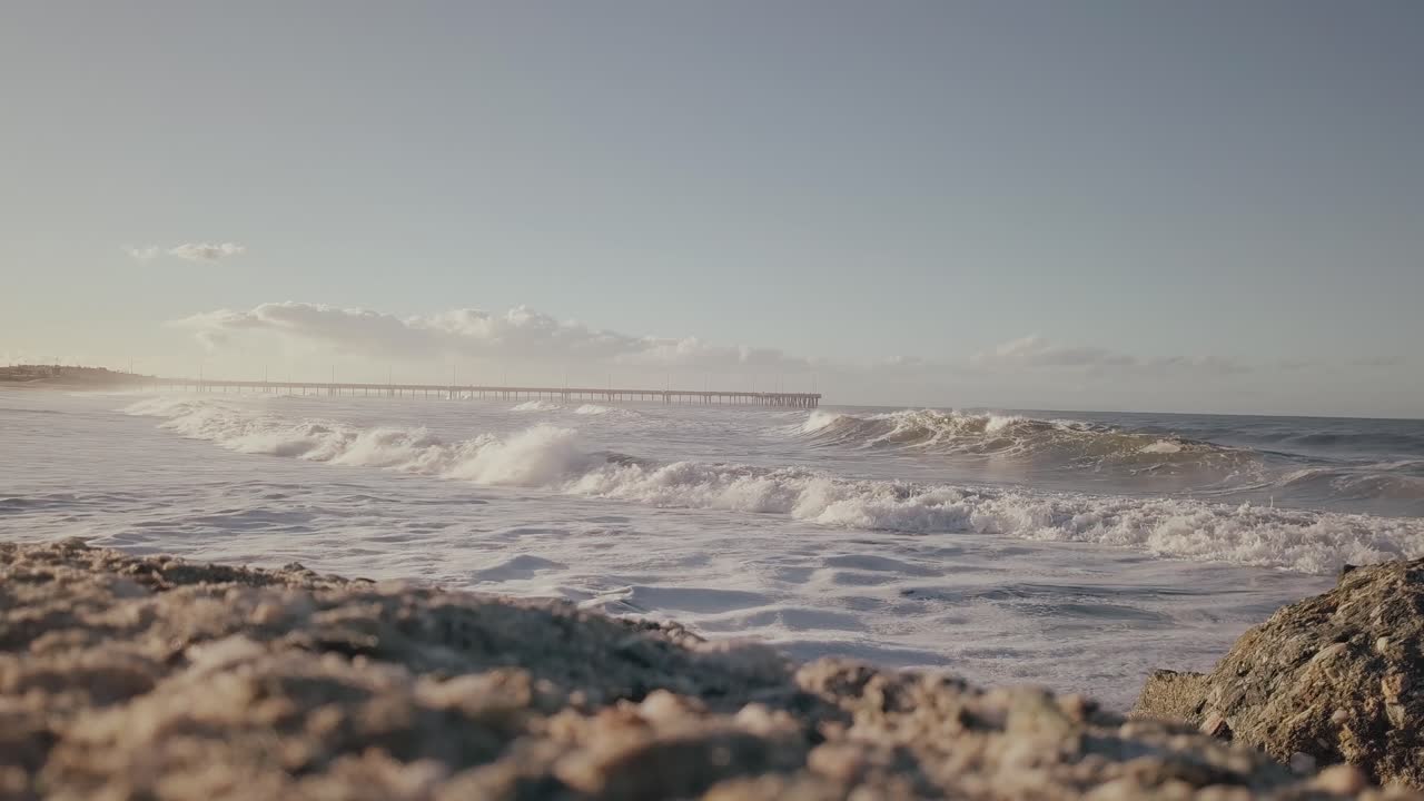 슬로우 모션 모래 해변을 닫고, 화창한 날 거리에서 베니스 해변 부두의 전망을 감상할 수 있는 롤링 파도