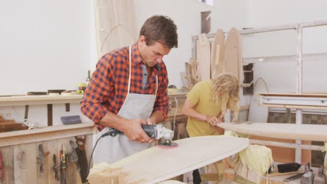 Two-Caucasian-male-surfboard-makers-working-in-their-studio-and-making-a-wooden-surfboard-together