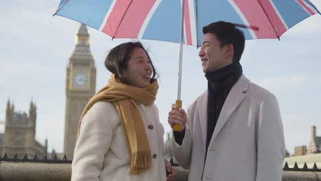 young asian couple on holiday posing for selfie in front of houses of parliament in london uk with umbrella 1