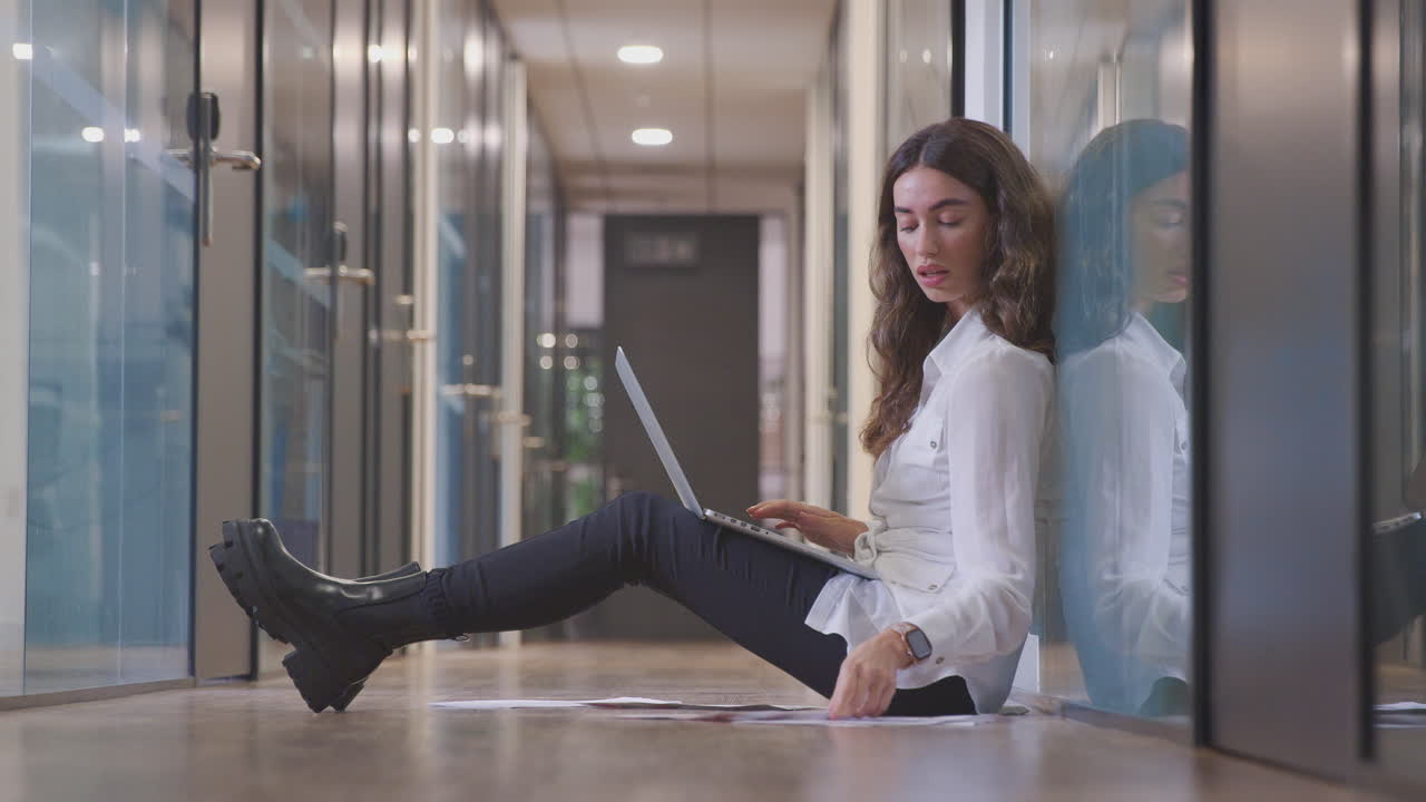 Giovane donna d'affari seduta per terra nel corridoio di un ufficio moderno che lavora al laptop.