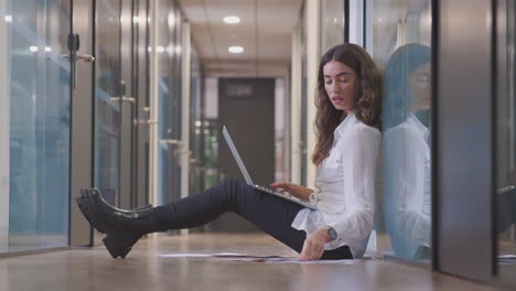 young businesswoman sitting on floor in corridor of modern office working on laptop