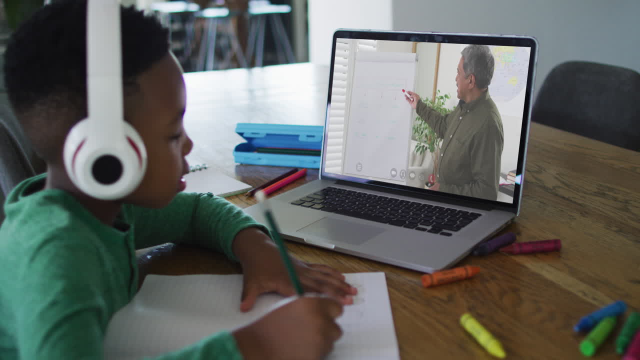 African american boy wearing headphones having a video call on laptop while doing homework at home