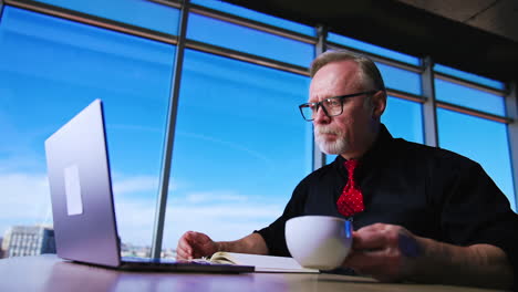 Old Caucasian male drinks coffee looking at laptop. Coffee break at the working place in office. Low angle view.