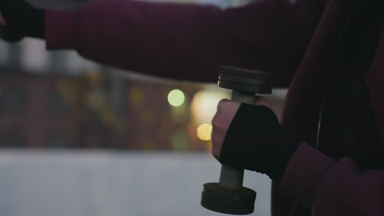 Close hand view of female in hoodie punching air with dumbbells, rapid boxing movement against urban blurry dusk background, wearing fingerless gloves, strength training on asphalt court by fence