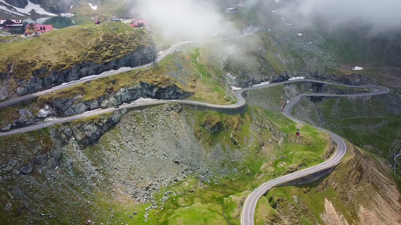 Drone aerial view over Transfăgărășan, winding road, Balea lake lodges, rocky cliffs and mountain mist.