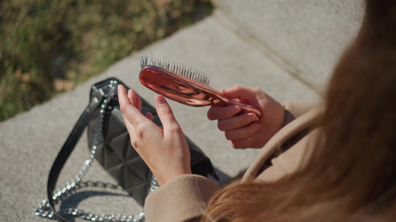 Primo piano di una donna che tiene ed esamina una spazzola arancione vicino alla sua borsa trapuntata nera su cemento con vegetazione sullo sfondo. Le mani del soggetto interagiscono delicatamente con la spazzola mettendone in mostra la.