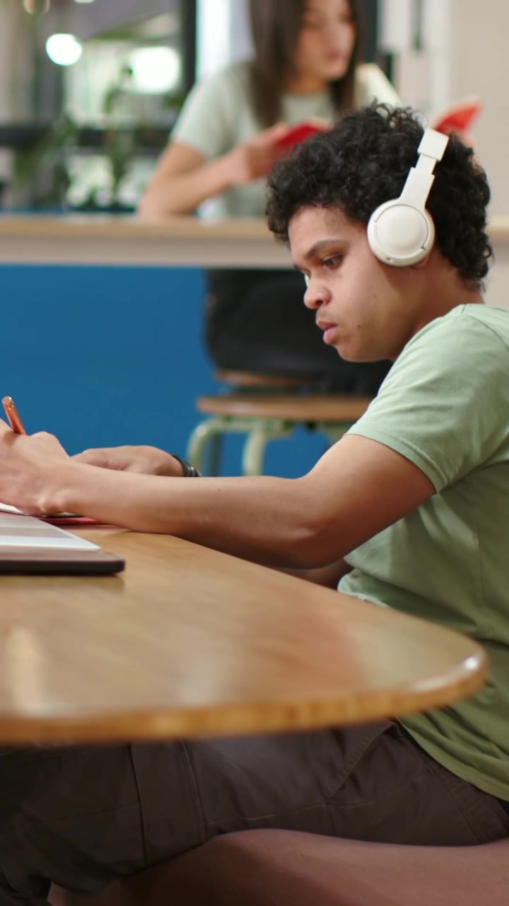 Vertical video: Starting lean, man typing laptop, taking notes at study table in green tee, headset