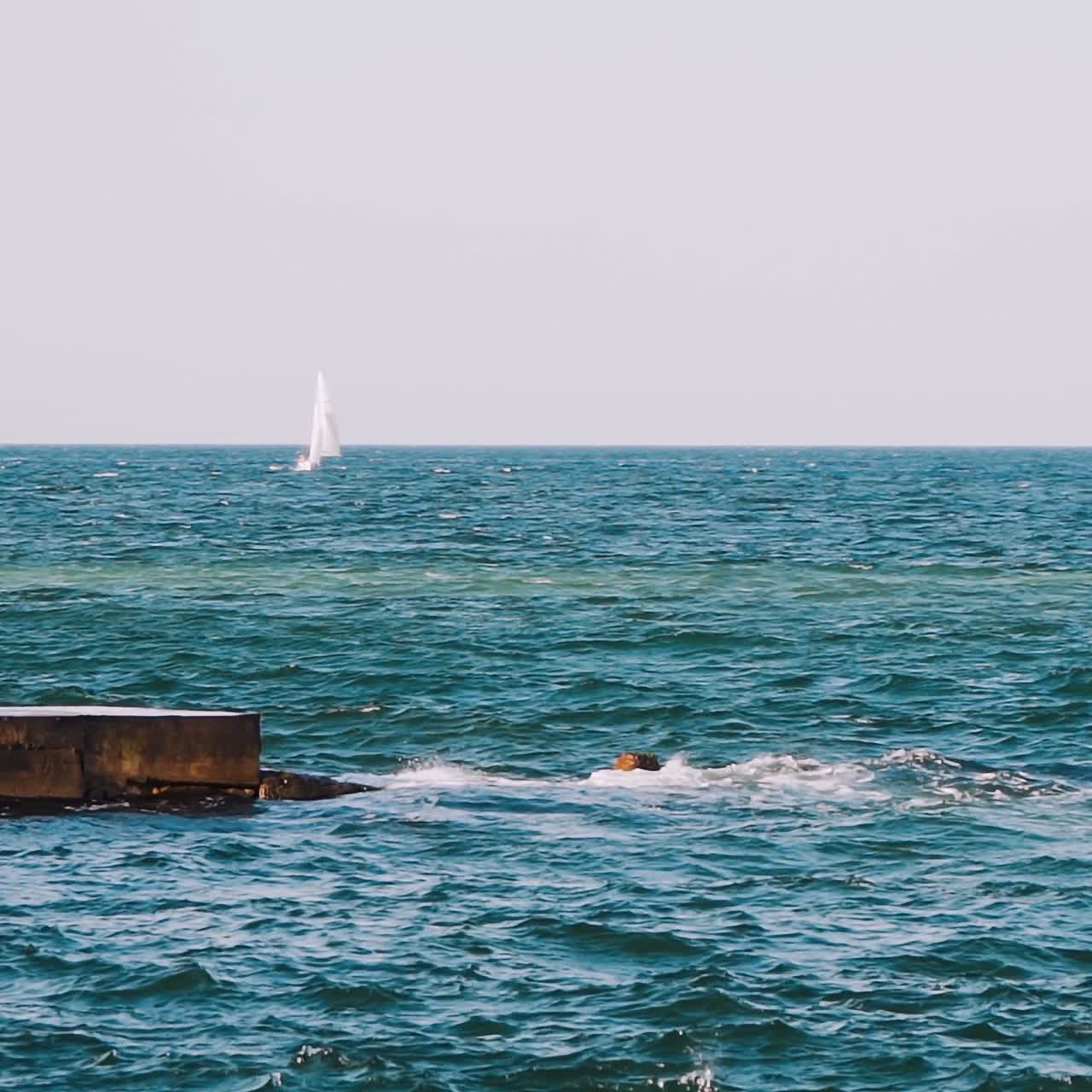 Motion of sea water. Waves break at stone pier and make white foam. Beautiful blue sea and white sail floating on alone. Slow motion.