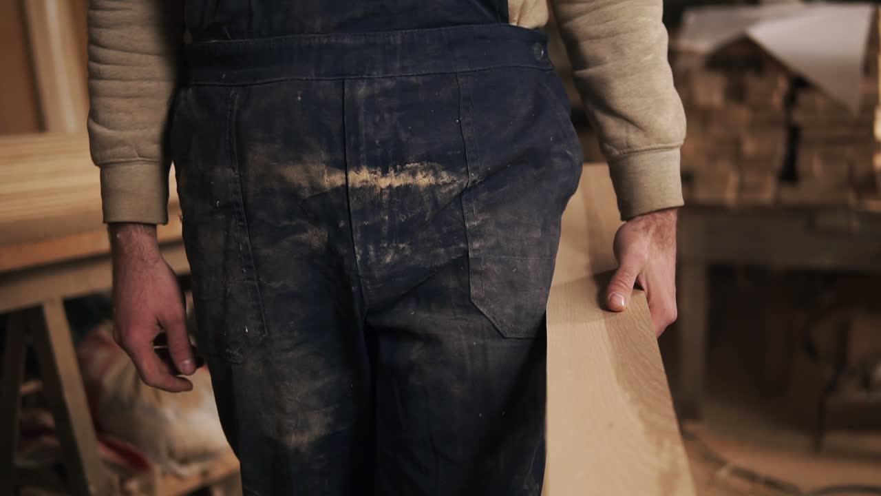 Slow motion footage of a young carpenter with rough hands. A man walks in a dirty with wood dust blue overalls with a bar of wood in his hands