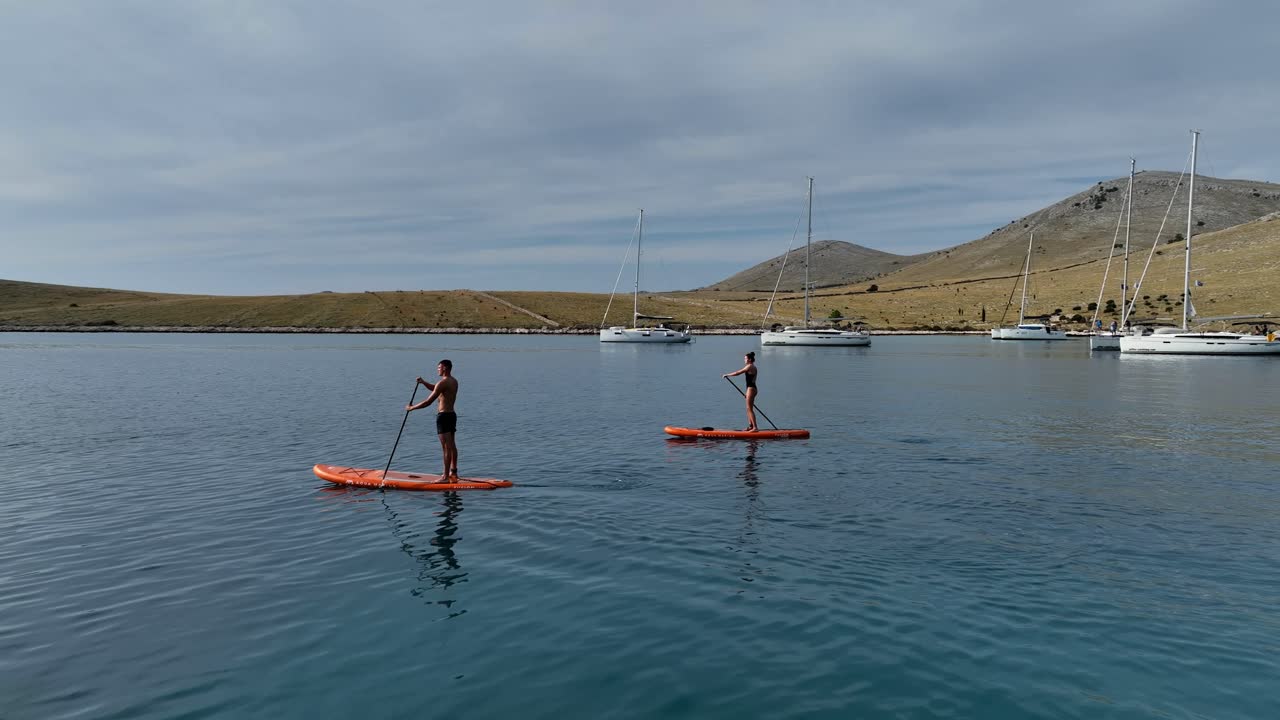 Two paddleboarders glide across calm waters with rolling hills in the background in Kornati, Croatia, medium aerial orbit