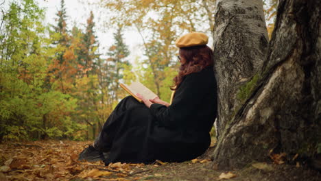 focused woman seated outdoors in autumn forest leans against tree, deeply engrossed in book, wearing yellow beret and black coat, she enjoys peaceful solitude surrounded by vibrant fall foliage