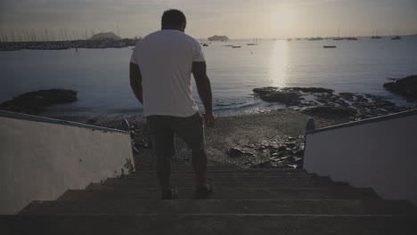 African-man-sits-on-staircase-facing-the-sea-at-sunrise