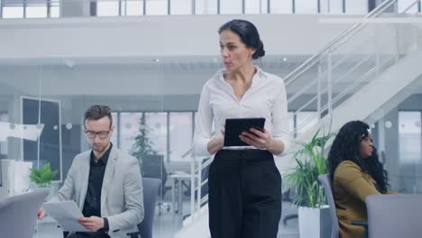 in the bright corporate office: female executive holding digital tablet computer walks through the office where her professional staff of businesspeople work on desktop computers