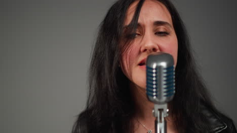 close view of a singer in a black leather jacket performing passionately with a vintage microphone against a grey background. the singer's expressive face and hand gestures