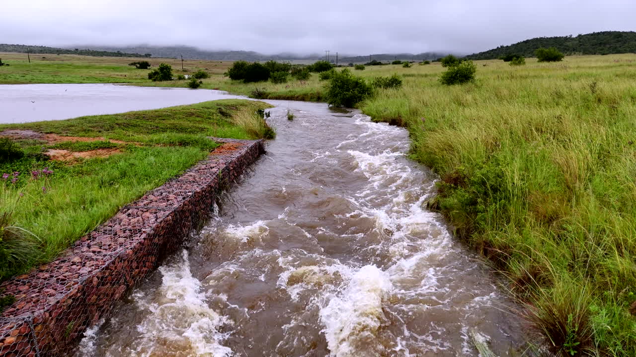 Rain floodwater overflowing from rural dam flooding past erosion barrier, aerial