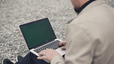 man is sitting on gravel beach and working with notebook with chroma key screen