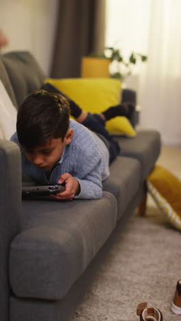 vídeo vertical de un niño acostado en el sofá en casa jugando a un juego o transmitiendo a un dispositivo de juego portátil 1