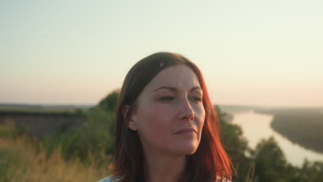 young woman with calm, pensive expression gazes to side in tall grassy field, golden sunset casting soft light on her face, with distant blurred river and treeline in background