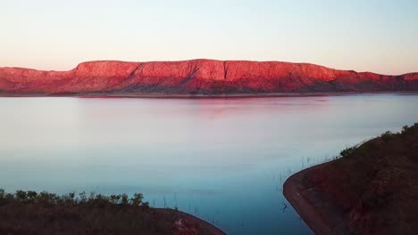 Straight,-fast-tracking-aerial-shot-of-dramatic-red-cliff-at-sunset-on-far-side-of-large-calm-lake-as-camera-tilts-up