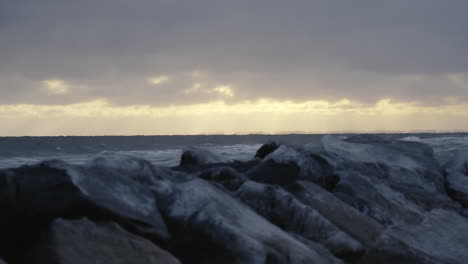 gentle waves with white crests gently wash against icy boulders as the sun begins to set on the horizon