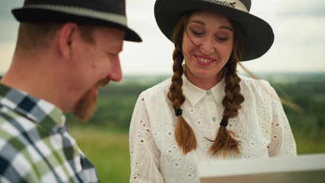 close-up of a joyful couple sharing a loving moment in a grassy field. the woman, dressed in white, opens a white box while laughing warmly with her spouse, who is wearing a black hat and plaid shirt