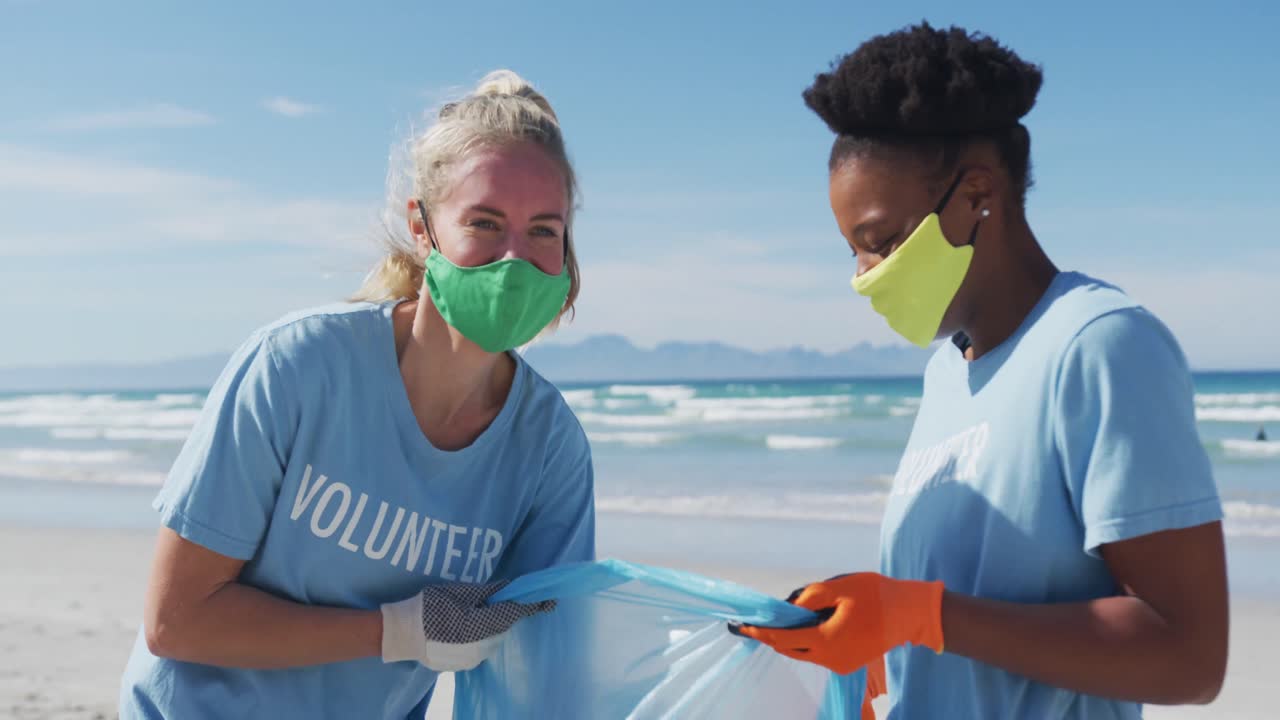 dos mujeres diversas con camisetas de voluntario y máscaras faciales recogiendo basura de la playa