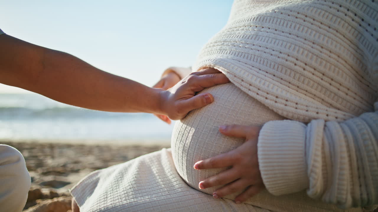 임신한 배를 만지는 남자의 손, 모래 해변에 앉아있는 아내, 가까운 미래의 부모