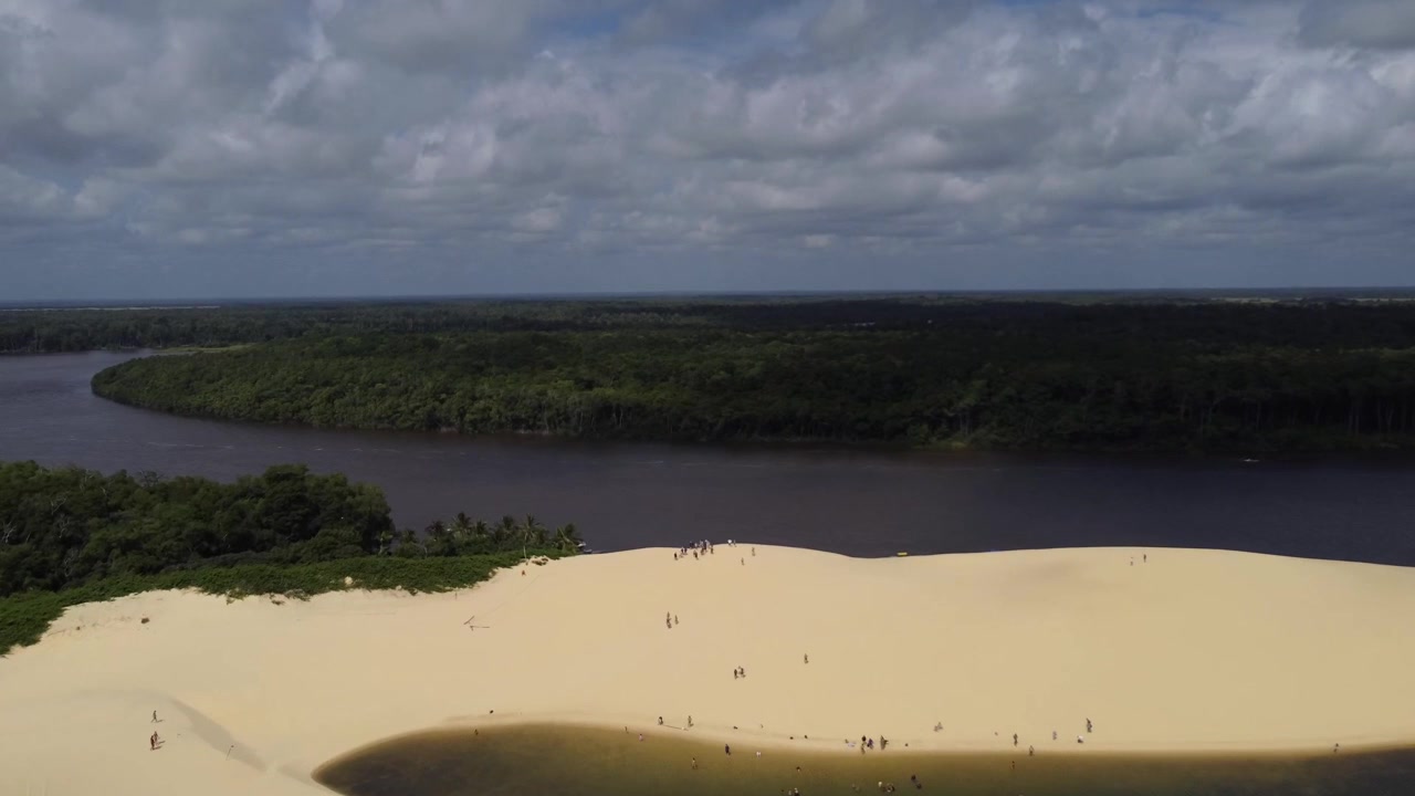 High-angle drone shot showing tourists walking on a large white sand dune with a freshwater lagoon and the wide Preguicas River in Barreirinhas, Brazil.