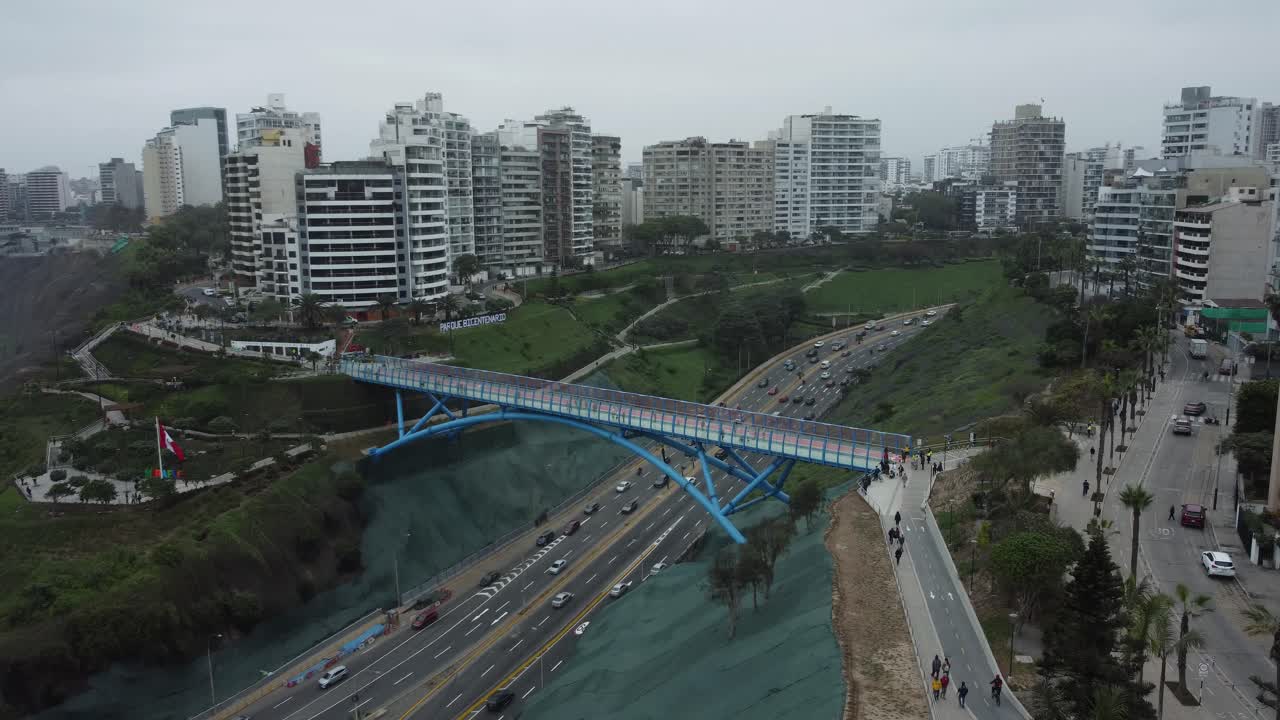 Aerial shot of a blue bridge connecting two districts of Barranco and Miraflores in Lima, Peru. Drone hovers in the air. Tall apartment buildings all around and below the bridge cars drive by and