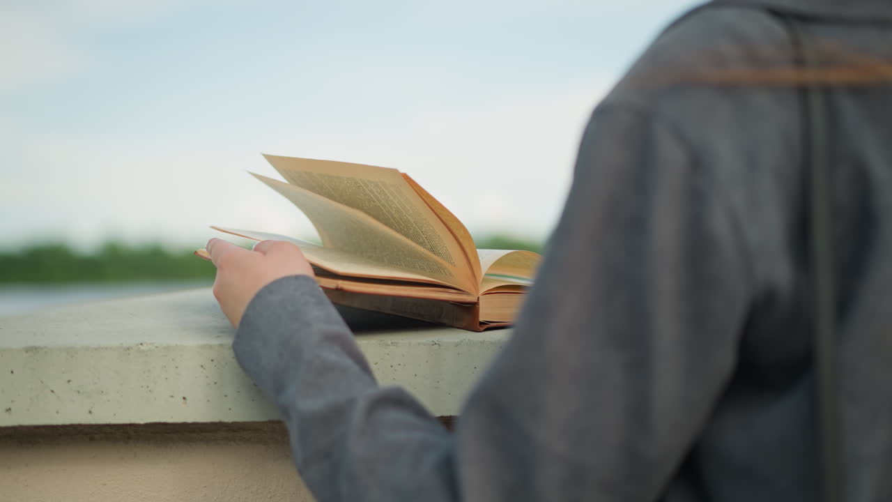 una dama con una camisa gris sosteniendo un libro en una valla, con el viento volviendo las páginas suavemente, ella está cerca de la tranquila orilla del río, con la vegetación visible en el fondo