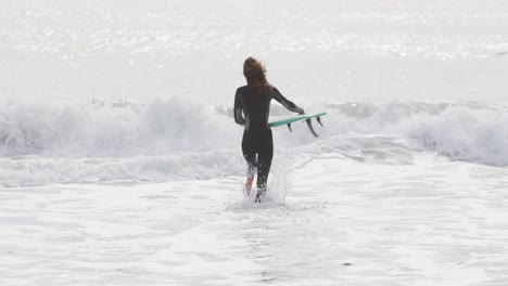 rear view of mixed race woman running into the sea carrying surfboard