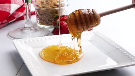 pouring sweet clover honey unto a white plate to rim a cocktail glass with fresh red strawberries in the background. making a strawberry shortcake martini with honey on the rim. pouring honey.