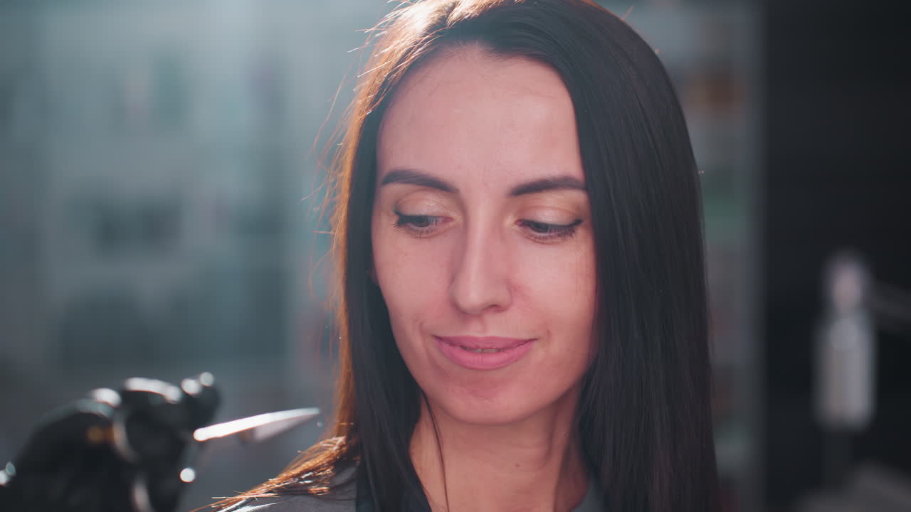 Mujer caucásica sonriendo en un salón de belleza tras un tratamiento capilar, con un cabello oscuro y elegante, suave contraluz que crea un halo, expresión de confianza y ambiente de estudio profesional con herramientas de estilismo desenfocadas al fondo.