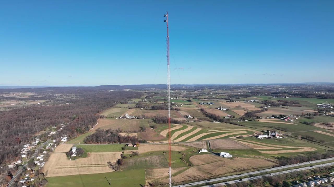 Rekaman orbit udara menara radio bergaris merah putih di daerah pedesaan kota Amerika. Lingkungan pinggiran kota dekat jalan raya dan ladang pertanian di musim gugur. Langit biru dan hari yang cerah. Sudut lebar