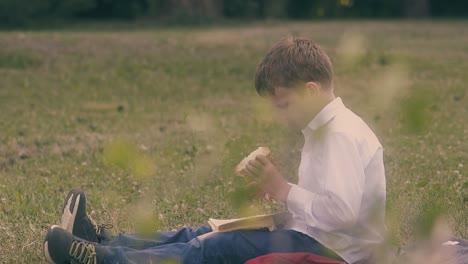 hungry boy with fair hair sits on lawn and eats big burger