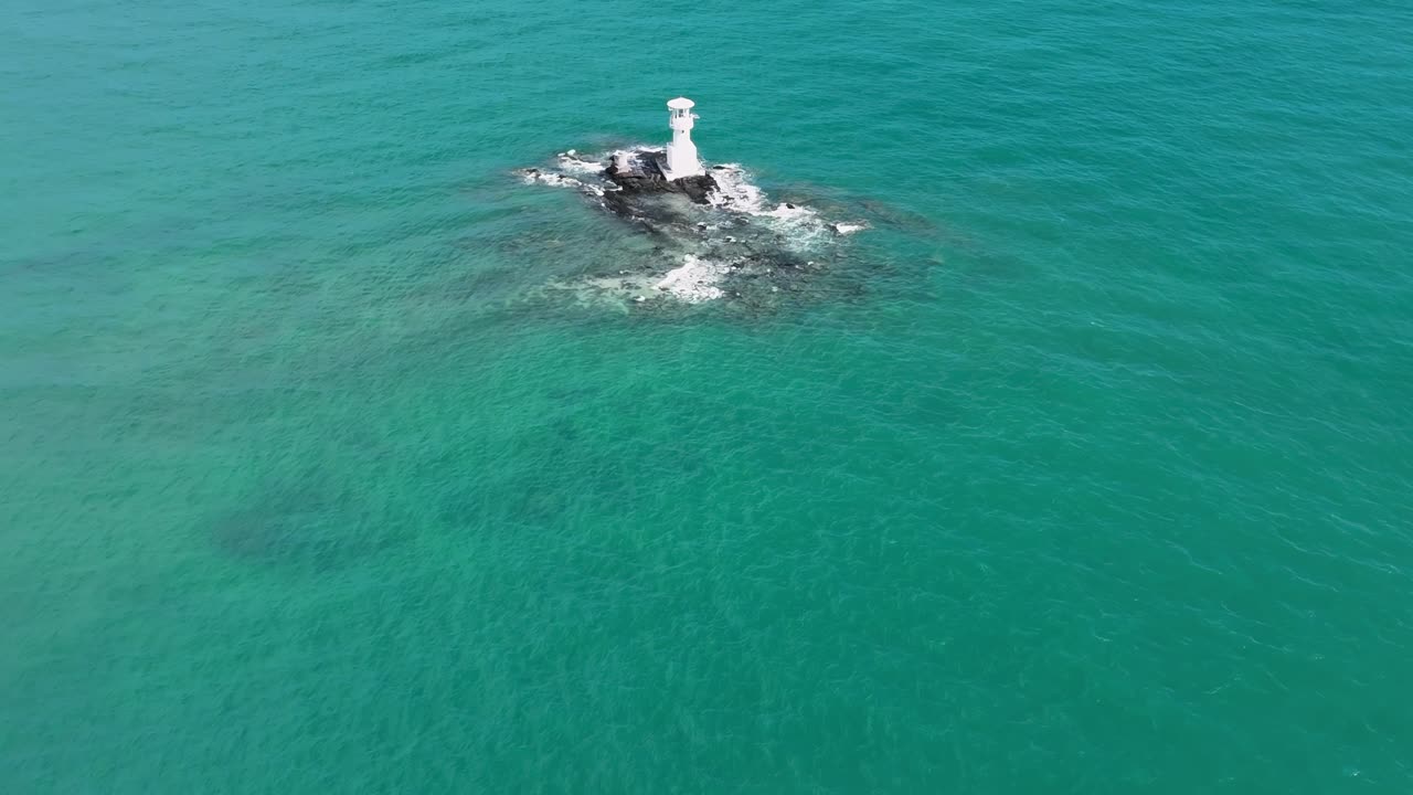 Aerial view of a solitary lighthouse perched on a small rocky islet surrounded by clear turquoise sea.