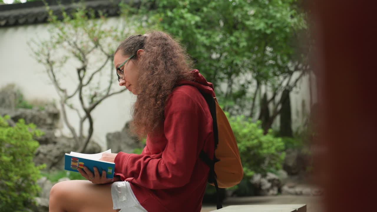 Ung student leser i hagen, krøllete hår og rød hettegenser, gul ryggsekk ved siden av, fokusert på notatbok og nettbrett, steinbenk og bonsaitrær i bakgrunnen, uskarp fotgjenger, mykt naturlig lys.