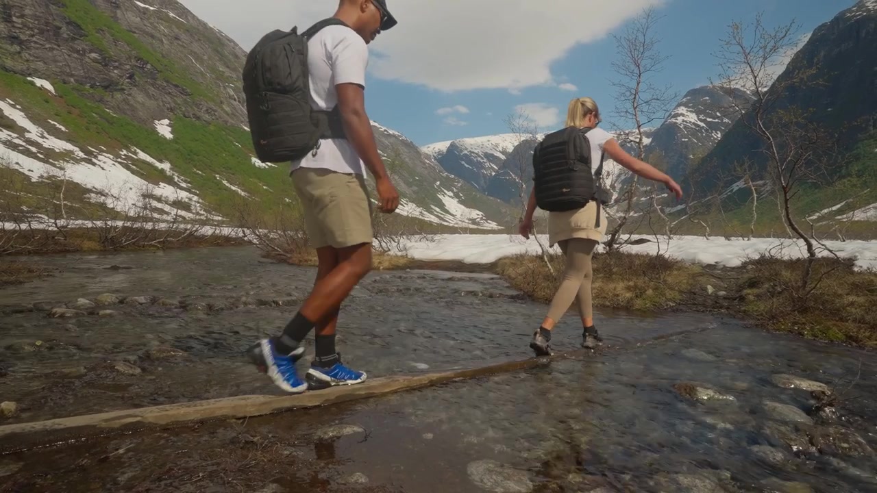Coppia di escursionisti con zaini che si bilanciano con attenzione su un piccolo ponte di tronchi di legno sopra un fiume poco profondo, circondati da montagne innevate nella Norvegia remota.