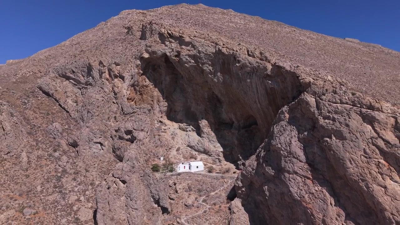 Ripresa con drone che rivela la cappella nascosta incastonata tra le rocce sopra la spiaggia di Perissa a Santorini, catturando le drammatiche scogliere vulcaniche