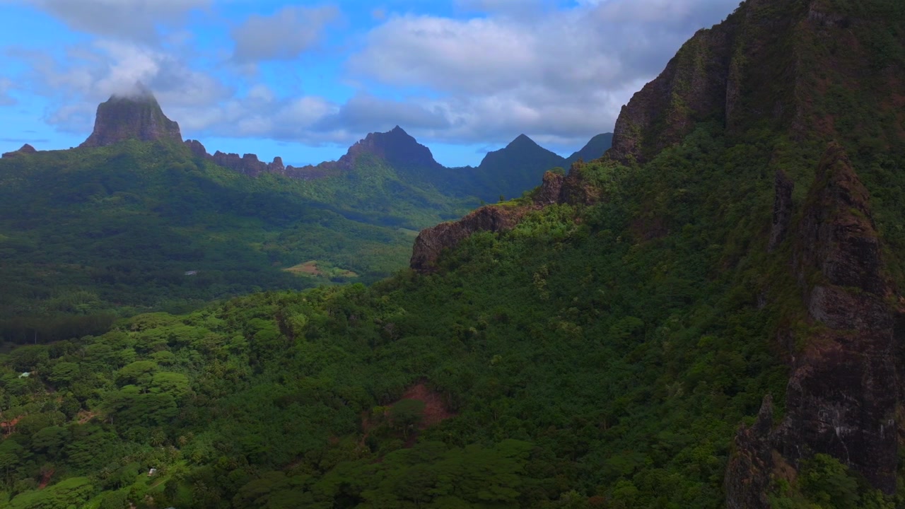 Dramatiska vulkaniska taggiga toppar, oländig terräng vid Mount Rotui och Mt Toheia, Moorea, Franska Polynesien. Flygfoto med drönare över Belvedere utsiktsplats, landskap över Cooks Opunohu-bukten, solig morgon, blå himmel, låga moln som cirklar åt höger.