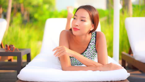 portrait of red-haired young thai woman resting lying on belly on deckchair leaning on elbows smiling and in hotel lounge in florida, usa - static slow-motion