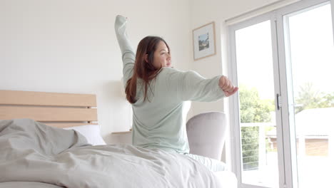 happy plus size biracial woman sitting on bed stretching in the morning, copy space, slow motion