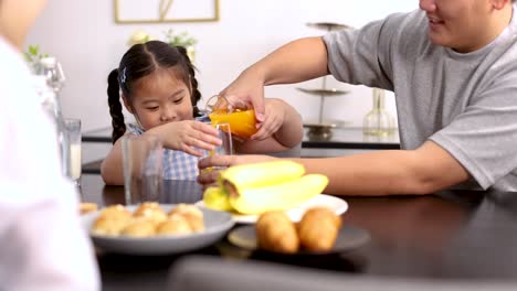 a father assists his young daughter in pouring orange juice at a breakfast table with pastries and fruit, in a bright, modern home setting
