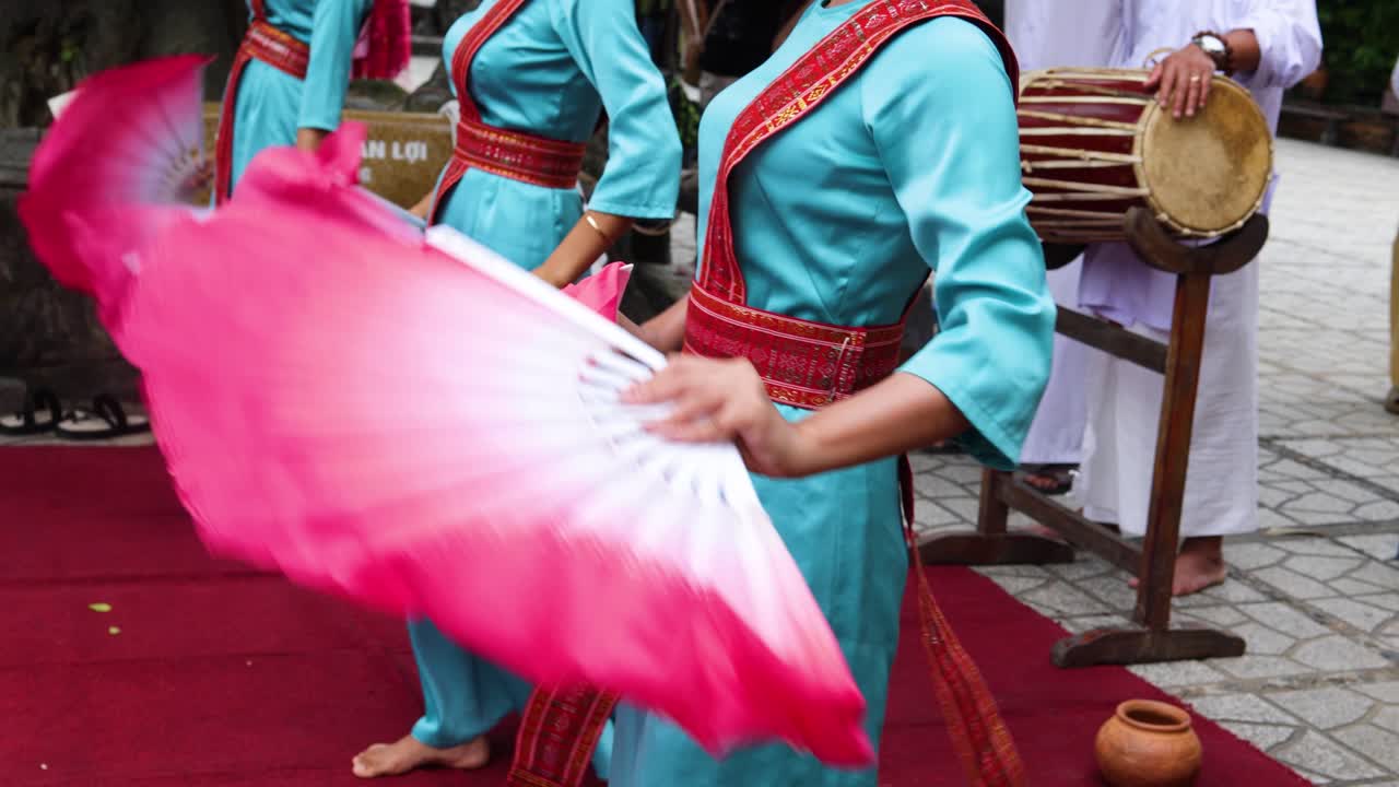 Penampil dengan pakaian adat pirus mengibaskan kipas pink besar selama upacara budaya di karpet merah di luar ruangan.