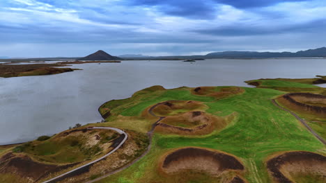 lakefront with craters of sleeping volcanoes, green grass and tourists paths nearby. grey cloudscape covering the sky above the scenery of iceland. silhouettes of mountains at backdrop.