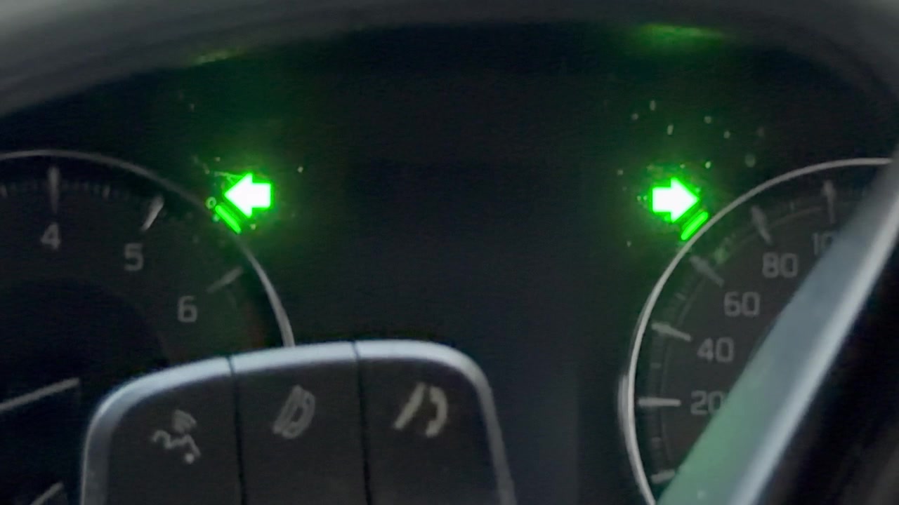 Close-up footage of a vehicle's instrument cluster at night, showing both left and right turn signal arrows illuminated in green as the hazard lights flash.